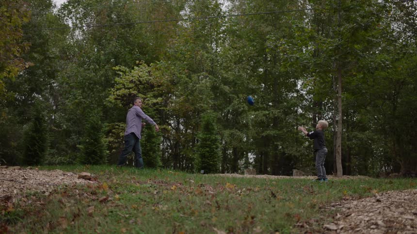 A man throws a football to his young grandson and the boy catches the ball and takes a step back as they play outside near the woods
