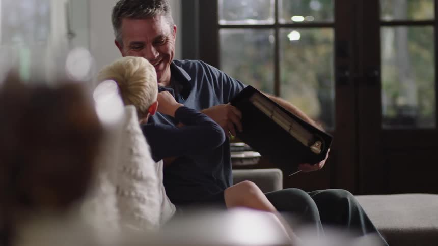 A man and child sit on the couch together as the man closes a book and the child makes faces at him