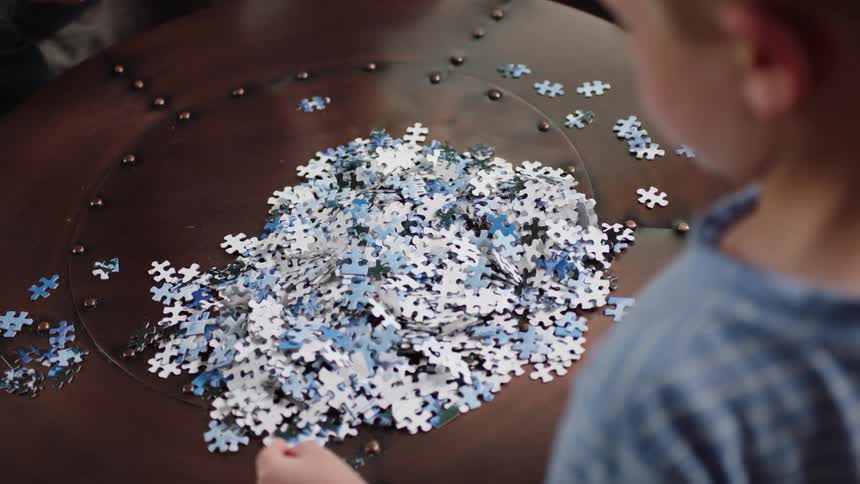 A little boy stands over a table looking at a pile of jigsaw puzzle pieces