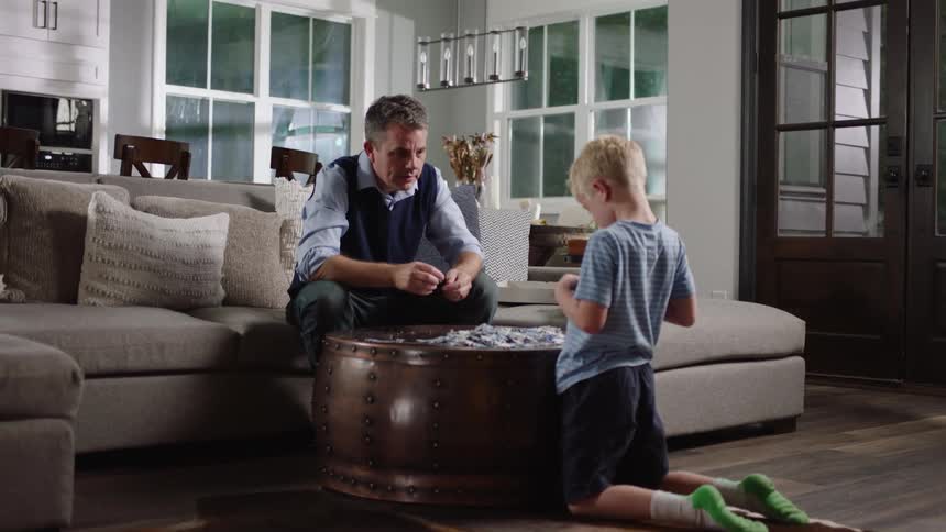 A little boy kneels over a table with a jigsaw puzzle as his grandfather hands him a piece of the puzzle