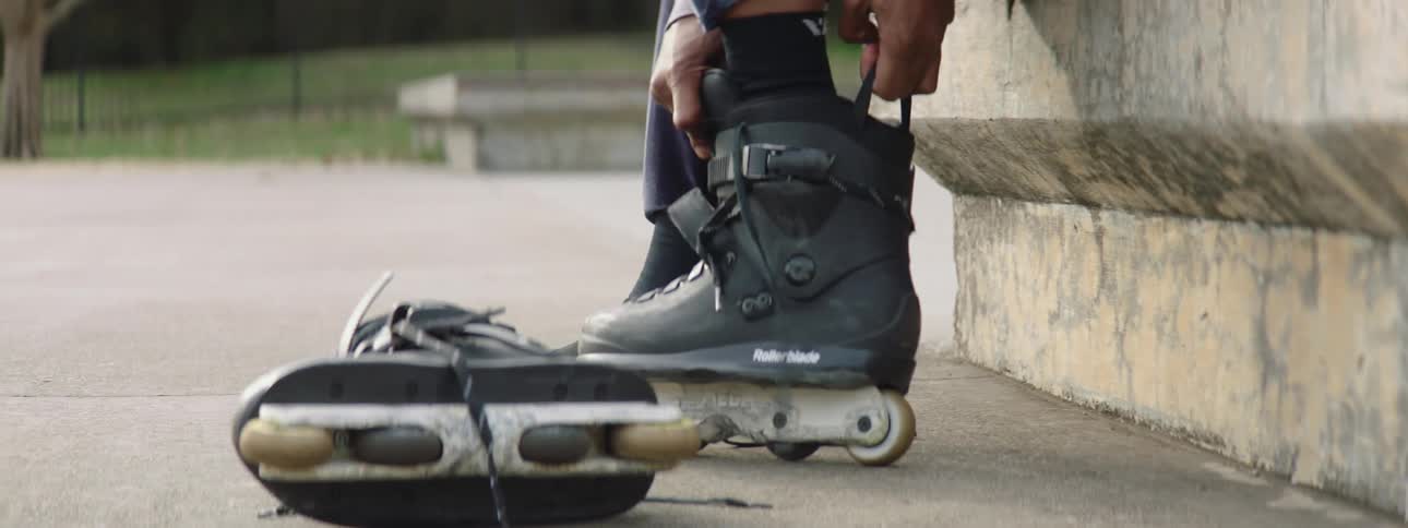 A young man is seated on a concrete bench at a skate park while putting on roller blade boots during the daytime