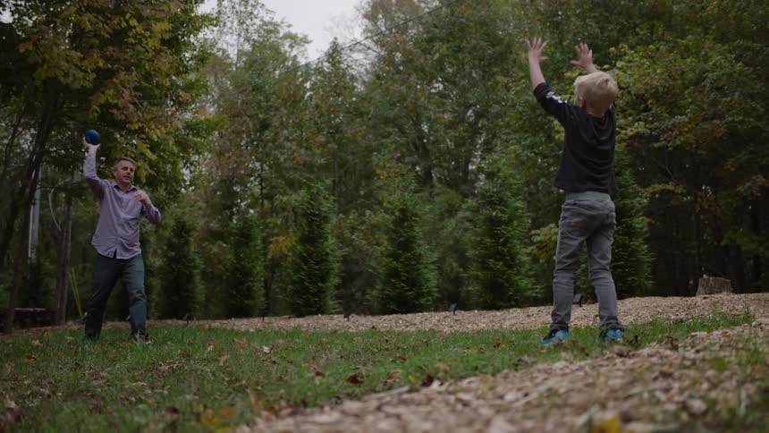 Father and son toss football together in park strewn with fallen leaves