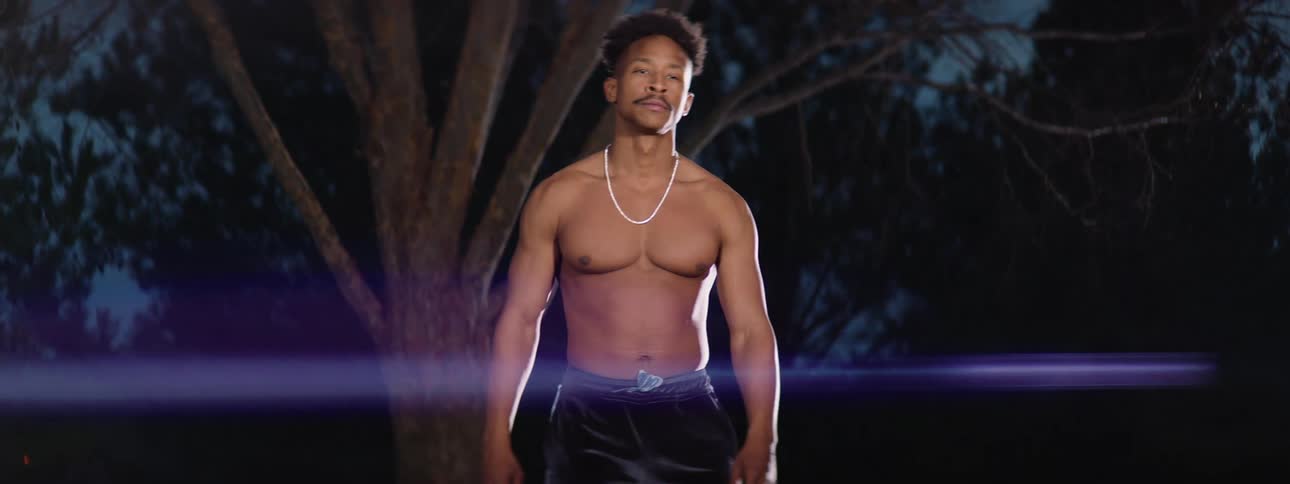Blue beam of light glares across scene as barechested man stands outdoors near trees