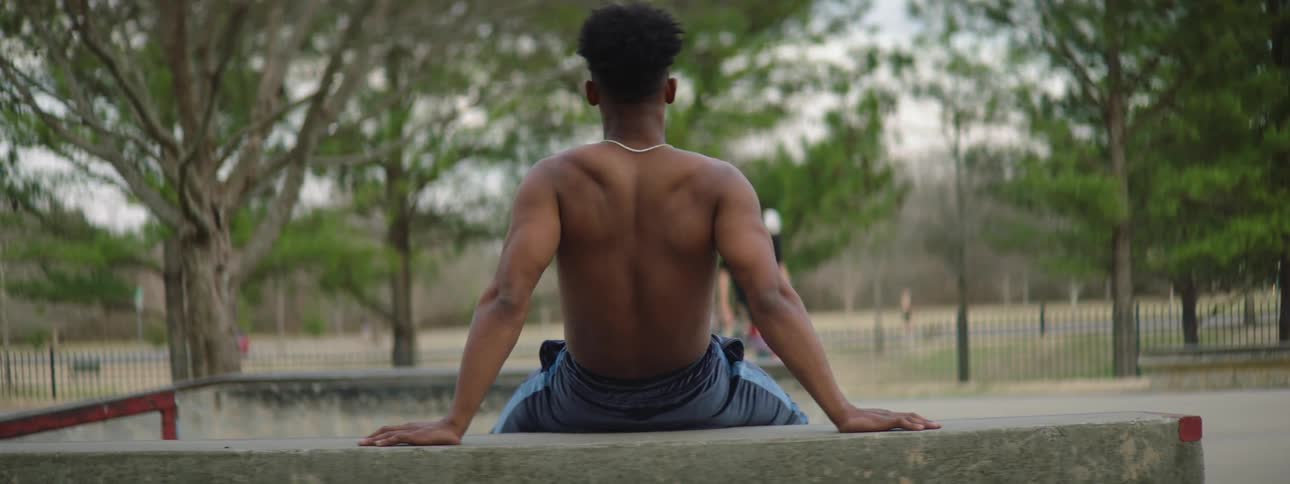 Back of a strong muscular shirtless man sitting on a park at daytime