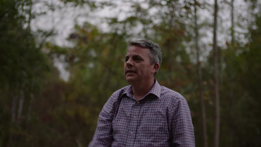 A man is standing outside near trees and woods with his hands up as he catches a football and gives a thumb up before pulling his arm back to throw the ball