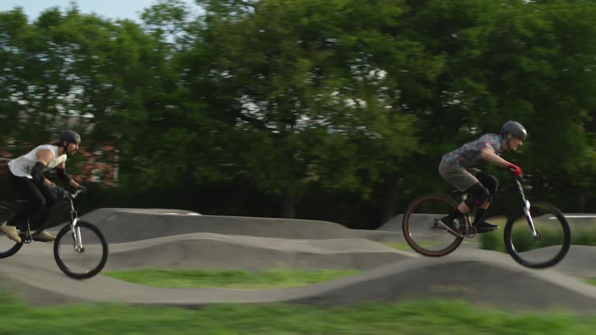 Two cyclists ride along ramps and slopes of park near housing