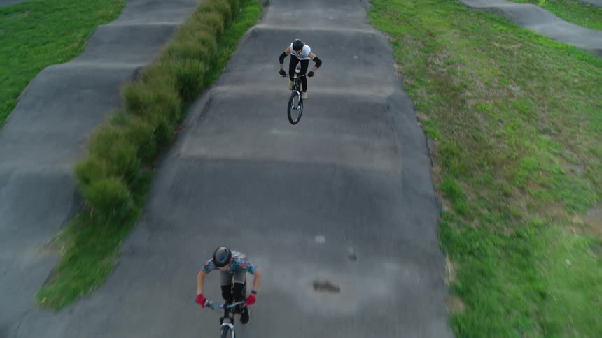 Two people are riding their bikes on a concrete path surrounded by grass and bushes and they do jumping tricks