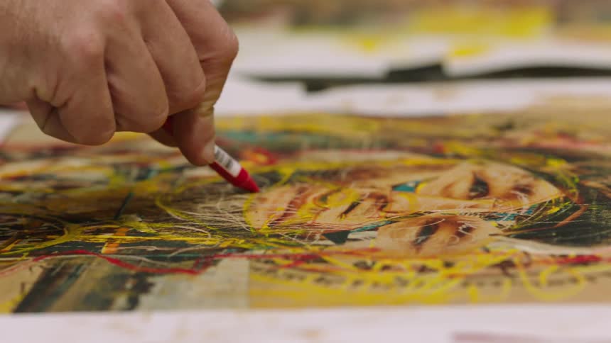 A mans hand is shown working on a drawing of a persons face with a red crayon