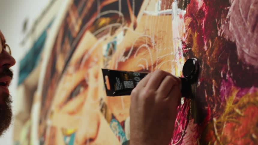 A man is adding the finishing touches of white paint being squeezed straight from the tube onto the already painted canvas in a graffiti style manner