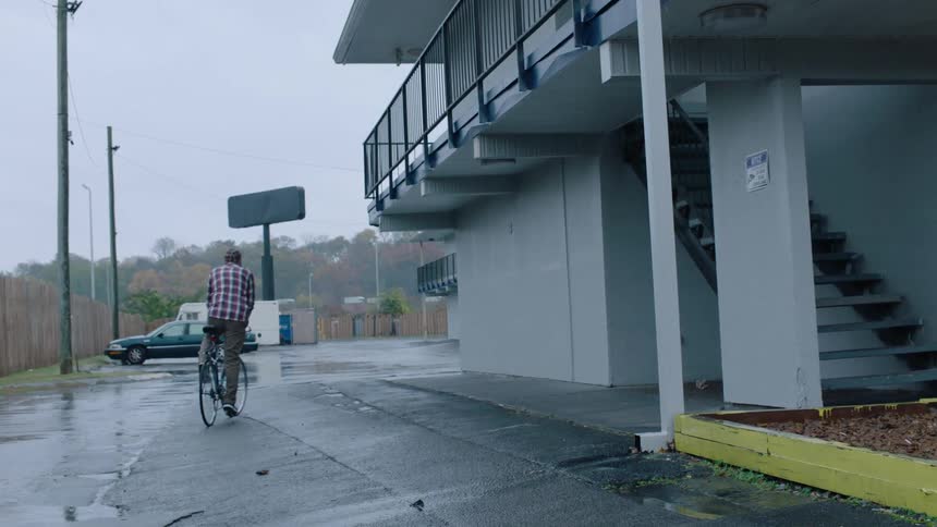 A man mounts a bicycle and rides it away from a building on a rainy day