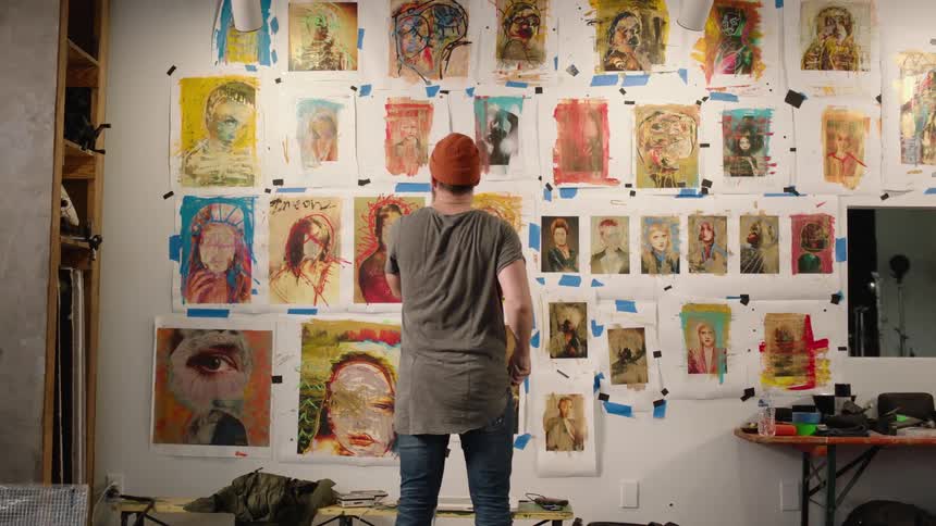 A man walks in slow motion up to a wall with several colorful abstract drawings taped to it and looks them over