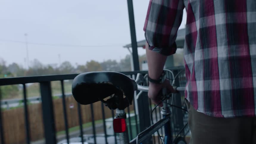 Man guides bicycle along corridor on outside of apartment building as vehicle moves along nearby road