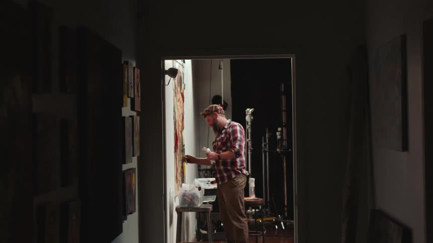An artist works on a painting using oil paintings in a studio at the end of a hallway