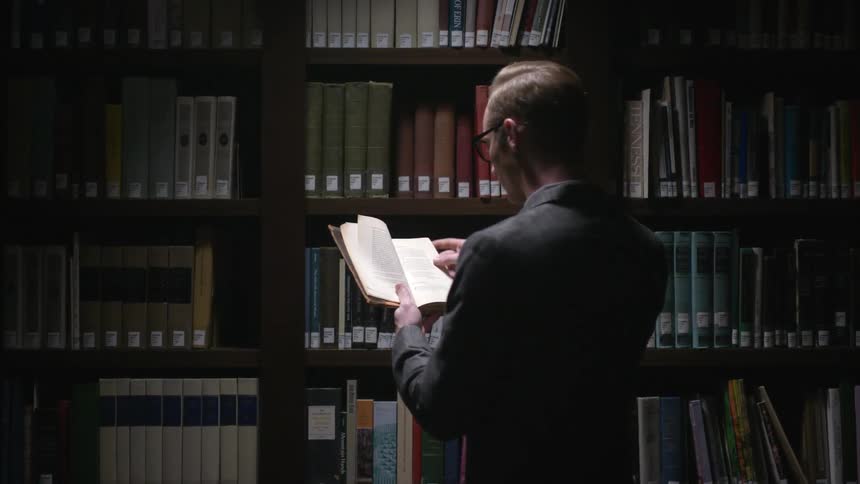 A man flips through a book in front of a bookshelf