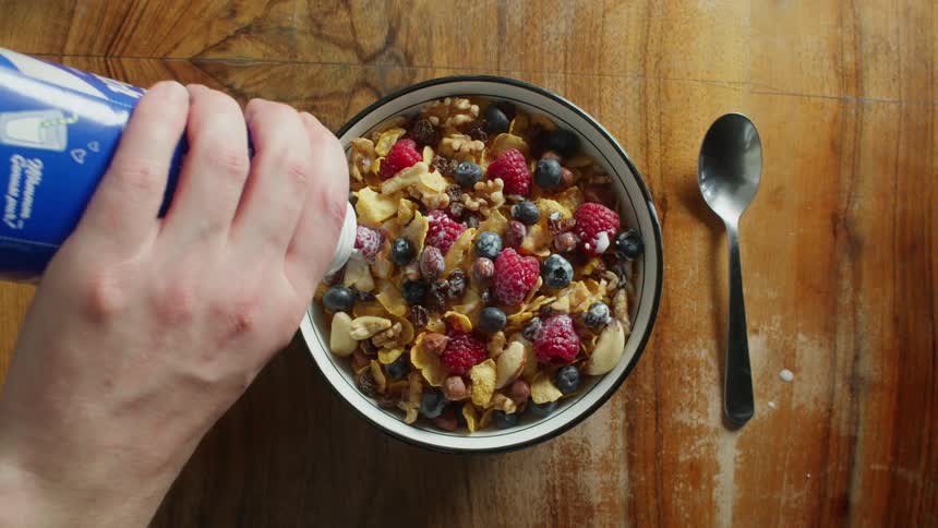 Pouring milk into a bowl of cornflakes with raspberries and blueberries on a table