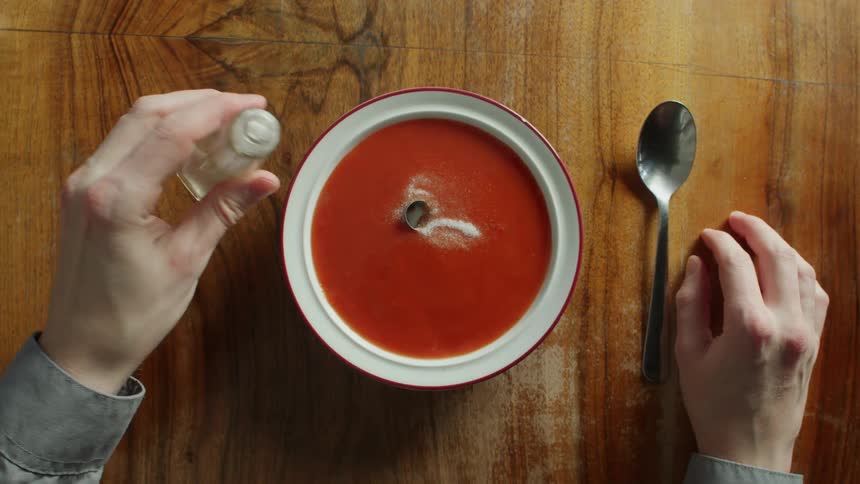 Pouring salt on a bowl of tomato soup on a table and the cap from the salt shaker comes off