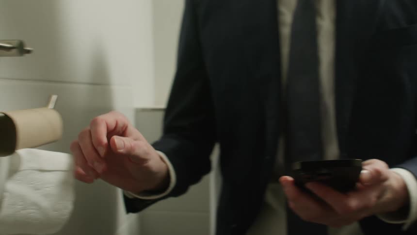 A man scrolls through his phone while in a public restroom stall with an empty toilet paper roll