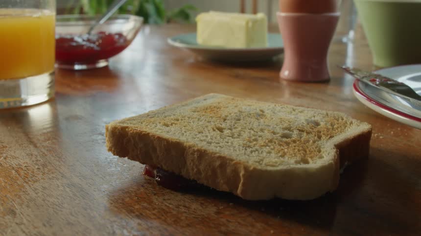 A man drops a piece of toast with jam face down on the breakfast table then picks it up leaving a mess
