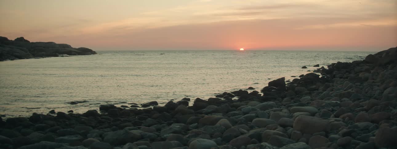 Sun sets over ocean as waves roll from red horizon toward dark pebble and rock covered shore