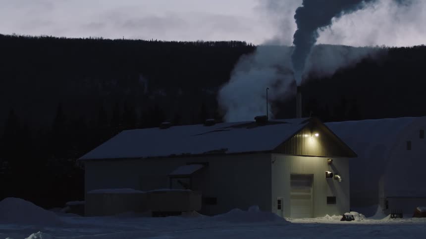 Smoke rises from chimney of business near woods after dusk during winter
