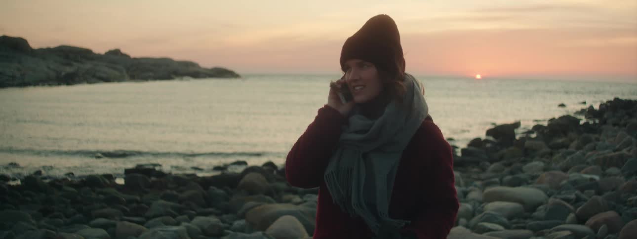 Woman with wooly hat gray scarf and coat talks on smartphone on the stony coast at sunset