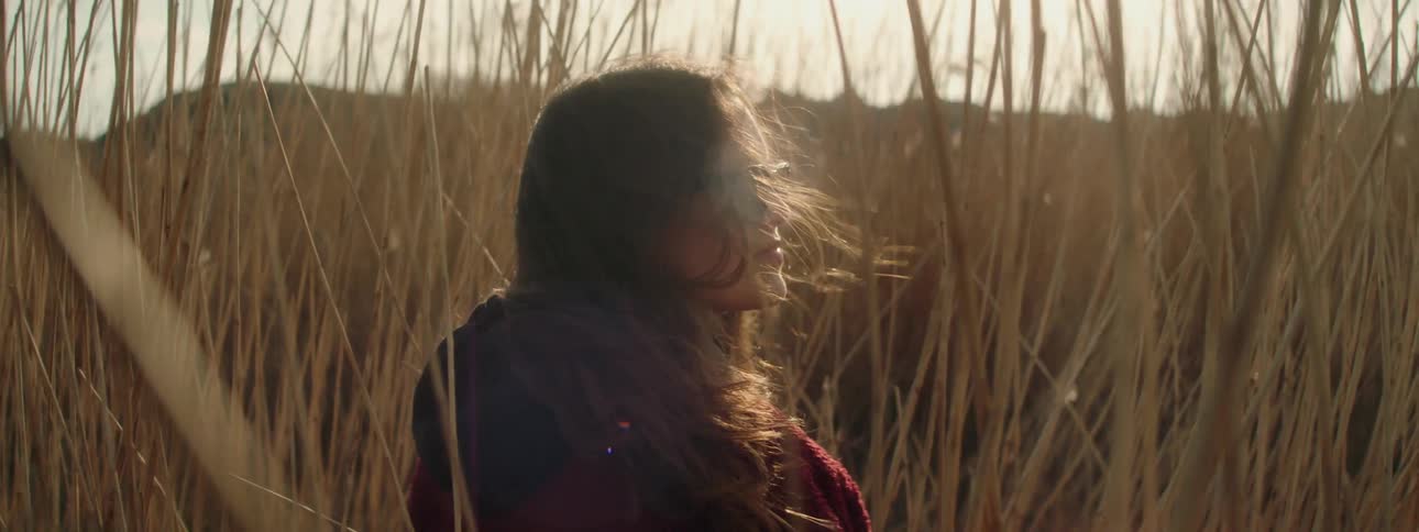 A woman wearing sunglasses is sitting outside in tall weeds as the wind blows her long brown hair