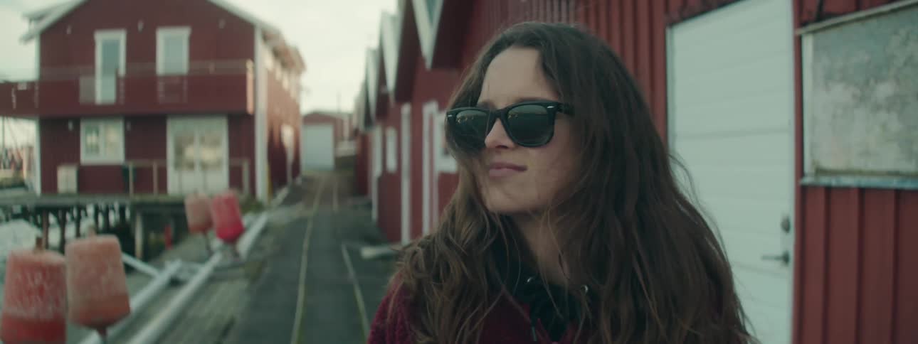 A womans face wearing black sunglasses with her hair blowing in the wind next to a dock