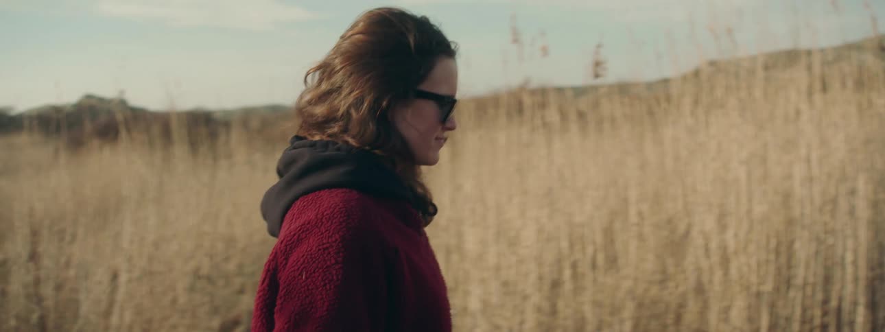 Young woman wearing sunglasses and scarf walking on a field of tall grass at evening