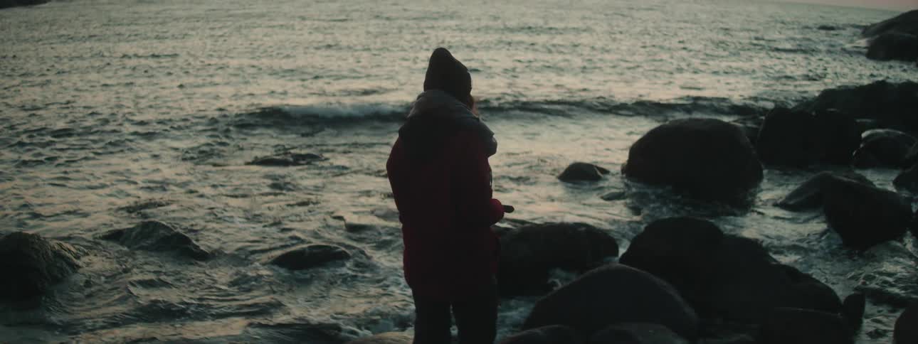 A woman wearing a scarf and hat stands near the coast line and rocks as waves crash up