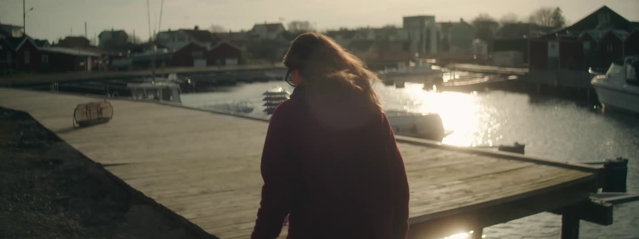 A woman walking with her hair blowing next to a wooden walkway with boats parked