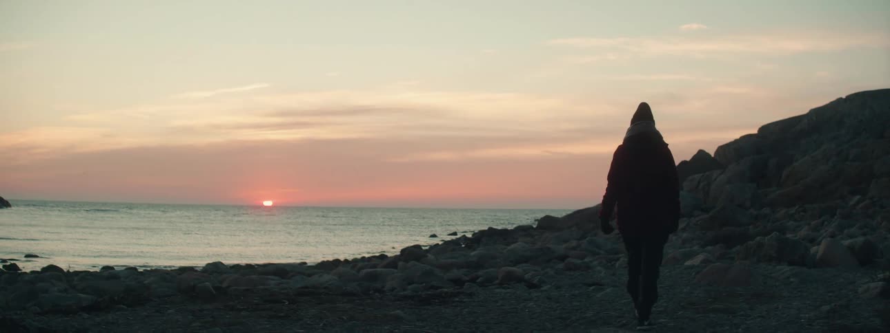 The sun is setting over the ocean as a person bundled head to toe in winter clothes a hat scarf and gloves walks along the edge of a rocky beach near the shore