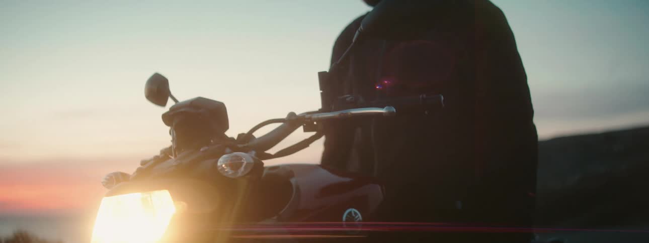 A man takes in the sunset along the coast while sitting on his parked motorcycle