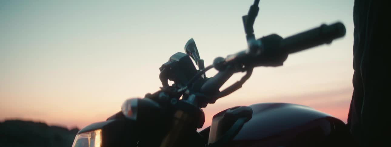 Man sits on motorcycle on hill over looking beach and ocean stretching to the red horizon