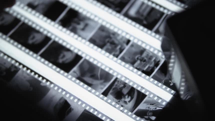 A persons hand holds a magnifying glass that looks over photo negatives in a dark room on a light board