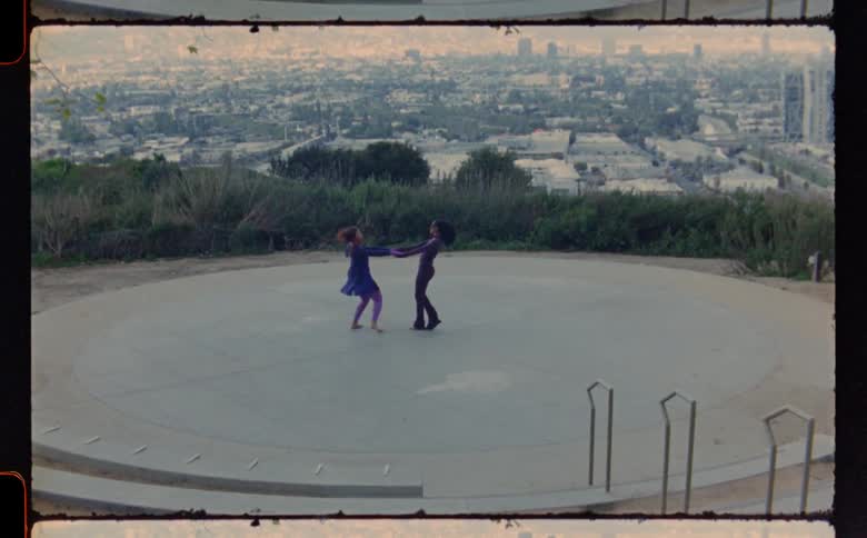 Two female dancers dance together on a circular plaza with city scape in the background