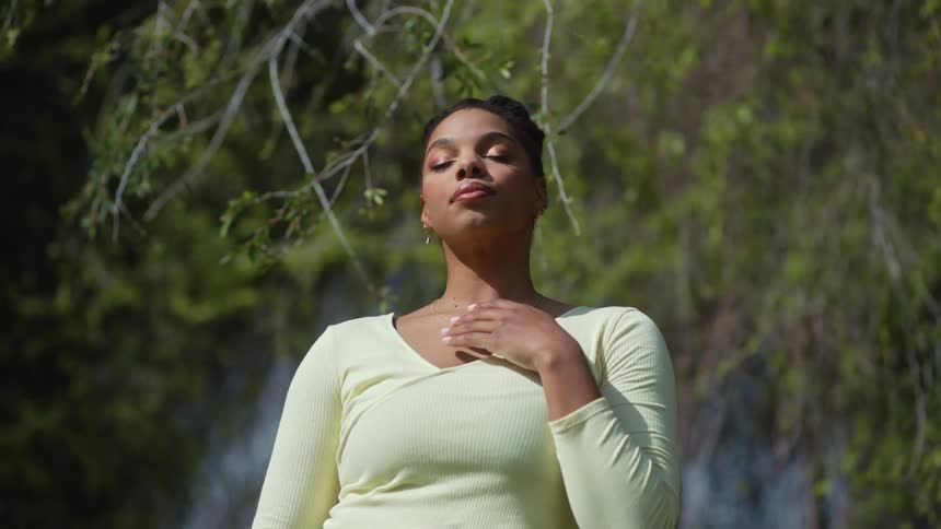 Woman runs her hand over her upper body while basking in sun near tree