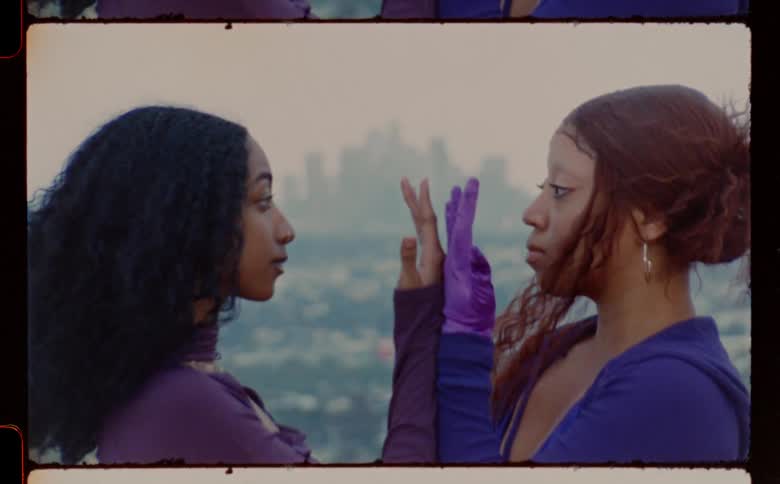Two women dressed in purple standing face to face in front of city skyline doing mirroring movements