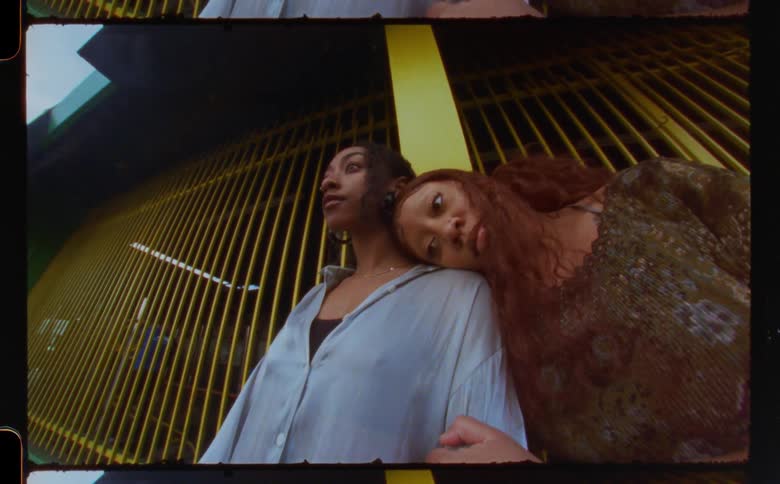 One woman leans on another as they stand near building with yellow bars