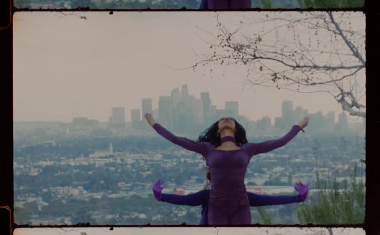Two black women wearing purple outfits moving their arms and hand in different ways one behind the other in a motion type dance with a city below them and a city skyline in the background