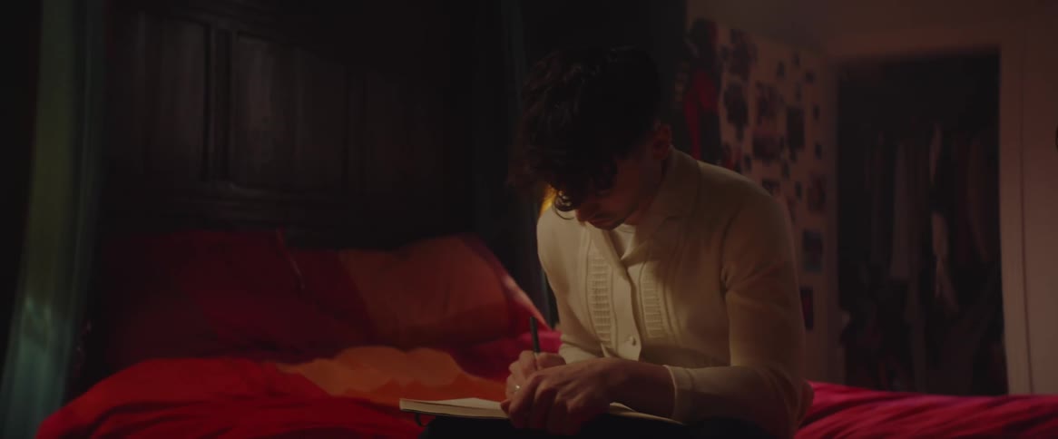 A teenage boy is sitting on his bed thinking as he writes in a notebook