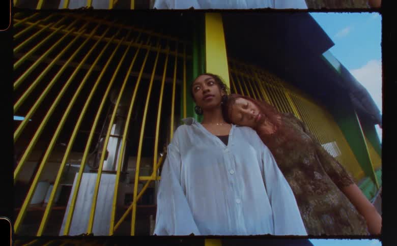 Two women standing against a yellow post with one woman resting her head on the others shoulder