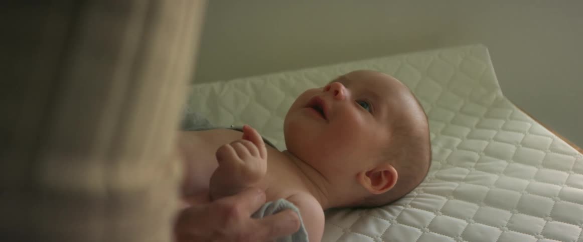 A newborn baby lays on a changing table while his mother plays with his hands and arms to amuse him