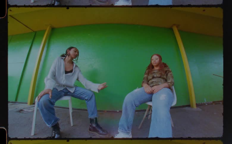 Two women sitting on chairs under overhang hold hands and smile