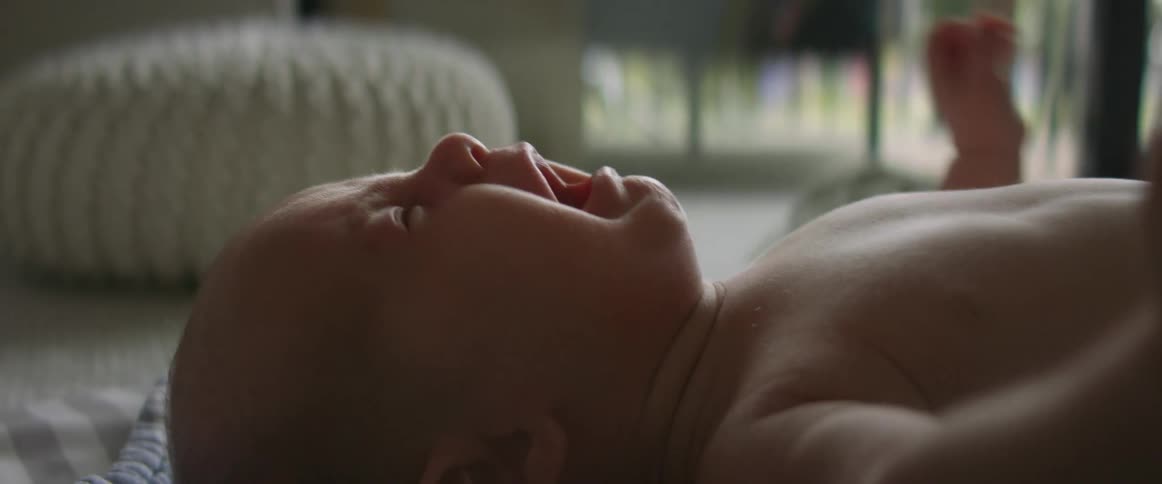 A shirtless cold baby is laying on their back with eyes squinted and mouth wide open screaming with arms are up and shaking around