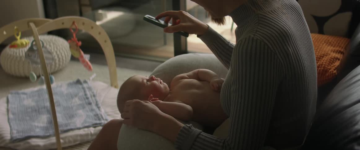 Blonde mother with sweater shows smartphone to baby son in the living room