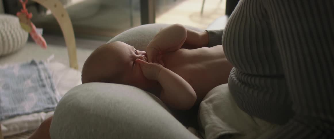 Mother touches and strokes baby lying on her lap and uses mobile phone sitting on couch in the living room