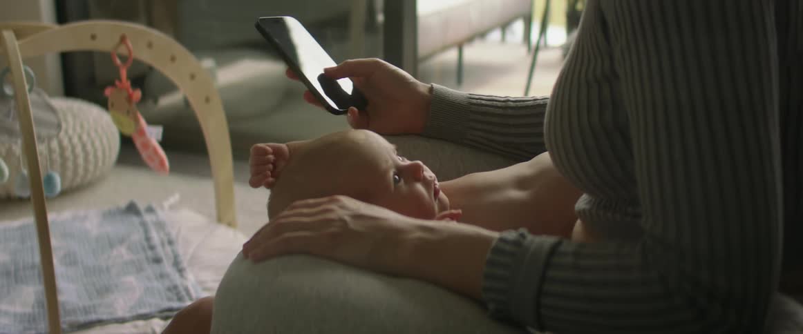 A mother holding her baby on a couch then picking up her phone and looking at it