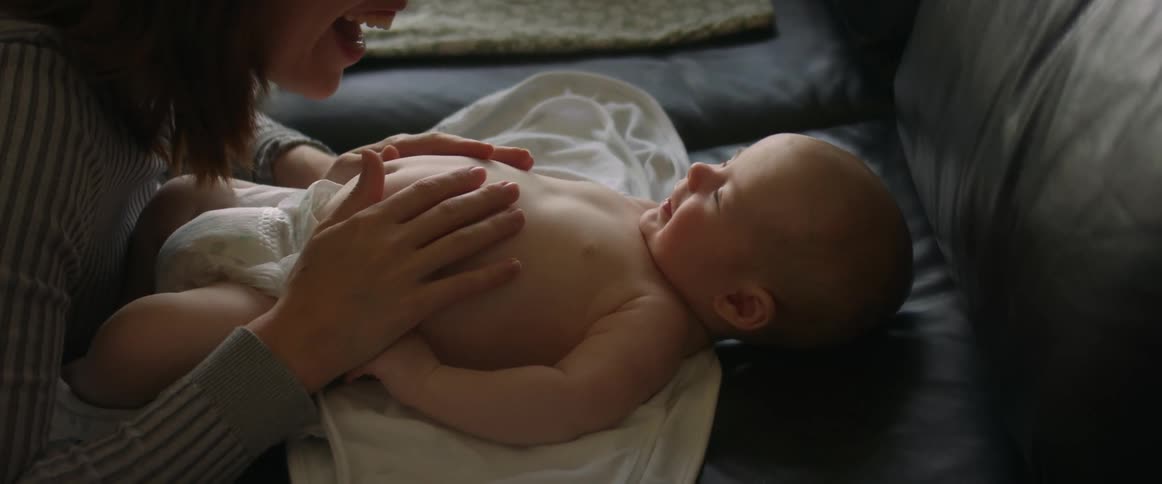 A baby in a diaper laying on a leather couch smiling at his mother making faces at him