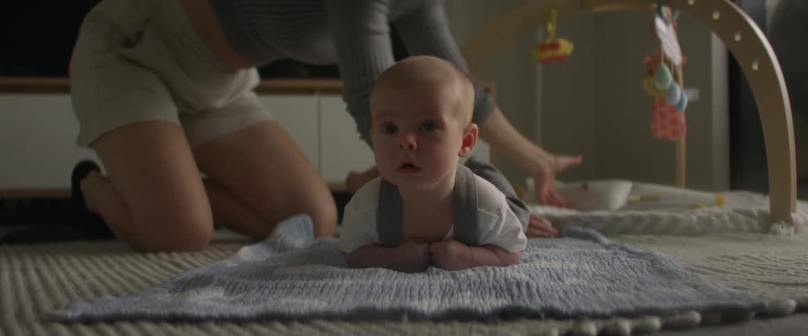 A baby is lying on a blanket on the floor as their mother picks up toys and straightens up the play area
