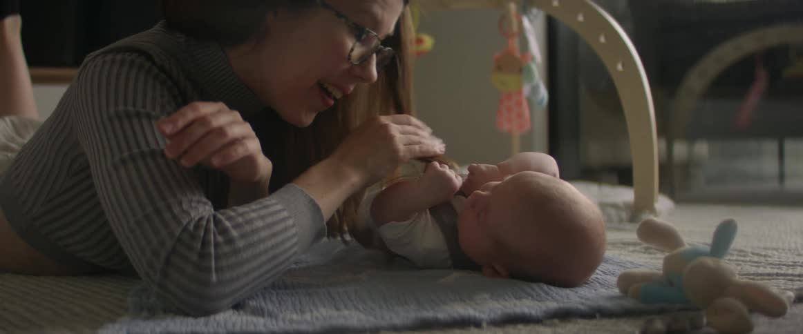 Mother with glasses and gray sweater looks at baby son lying down on the floor